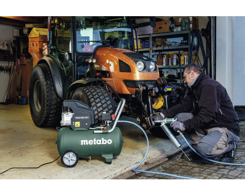 Eine Person nutzt eine Fettpresse an einem Metabo Kompressor Basic 250-50 zur Wartung eines Traktors in einer hellen Werkstatt.