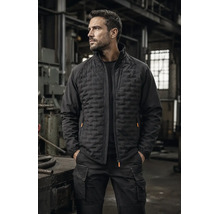 Mann in einer Werkstatt trägt eine schwarze Hybrid-Arbeitsjacke mit gesteppter Vorderseite und eine schwarze Cargo-Arbeitshose.