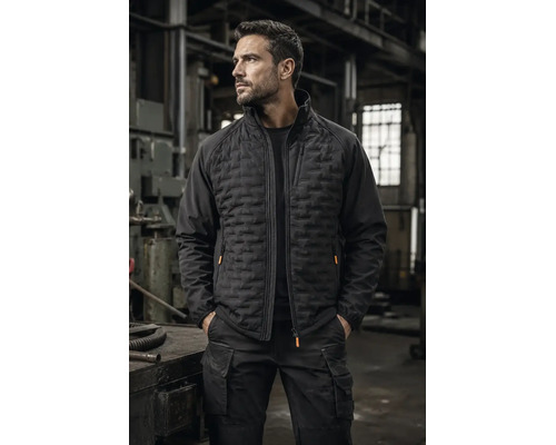 Mann in einer Werkstatt trägt eine schwarze Hybrid-Arbeitsjacke mit gesteppter Vorderseite und eine schwarze Cargo-Arbeitshose.