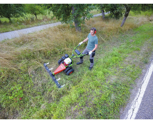 Ein Mann bedient einen Balkenmäher beim Mähen von hohem Gras auf einer Wiese am Wegesrand.