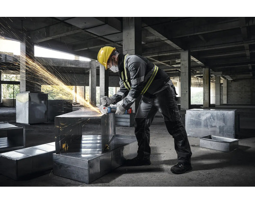 Handwerker in grauer Arbeitskleidung mit Winkelschleifer auf einer Baustelle bei der Bearbeitung von Metallkanälen.