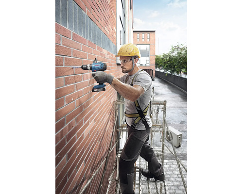 Handwerker bohrt mit Bosch Akku-Schlagbohrschrauber auf einem Gerüst in eine Ziegelwand. Er trägt Helm, Schutzbrille und Sicherheitsgurt.
