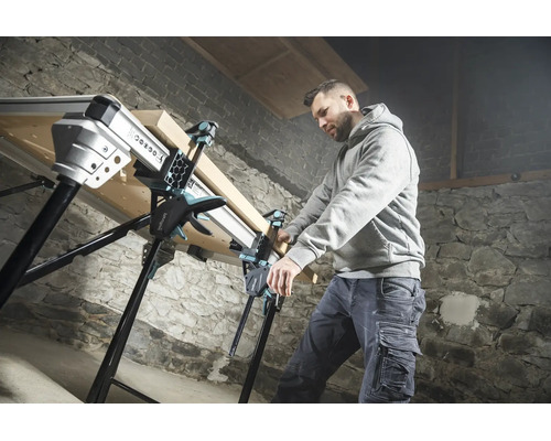 Handwerker fixiert ein Holzbrett mit Einhandzwingen auf einer multifunktionalen Werkbank in einer Werkstatt mit Steinwand.
