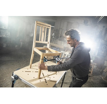 Handwerker repariert einen Holzstuhl auf einem Spanntisch mit einer Einhandzwinge in einer hellen Werkstatt.