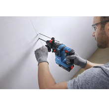 Ein Mann bohrt mit einem Akku-Bohrhammer von Bosch Professional in eine markierte weiße Wand. Er trägt Schutzbrille und Arbeitshandschuhe.