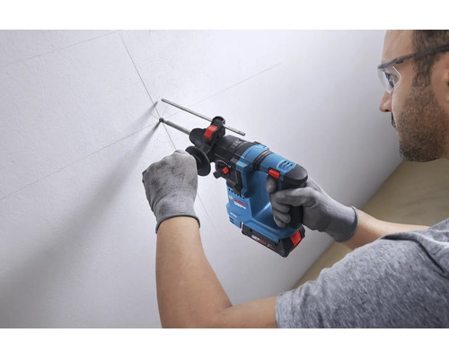 Ein Mann bohrt mit einem Akku-Bohrhammer von Bosch Professional in eine markierte weiße Wand. Er trägt Schutzbrille und Arbeitshandschuhe.
