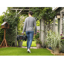 Frau trägt ein 18 Volt Akku-Gartengerät über einen grünen Rasen in einem gepflegten Garten mit Pergola und Pflanzen.