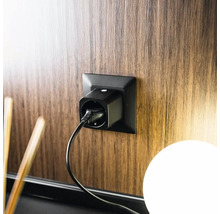Schwarzer WLAN-Zwischenstecker in einer schwarzen Steckdose an einer Wand in Holzoptik mit eingestecktem Kabel.