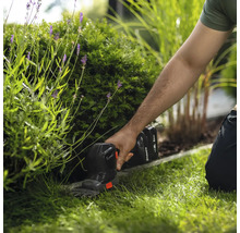 Akku-Grasschere von Husqvarna beim Schneiden einer Rasenkante im Garten neben einem Strauch.