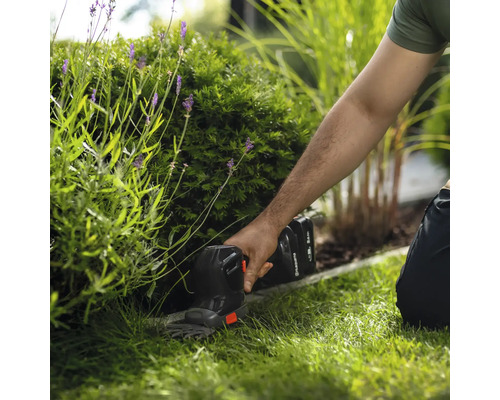Akku-Grasschere von Husqvarna beim Schneiden einer Rasenkante im Garten neben einem Strauch.