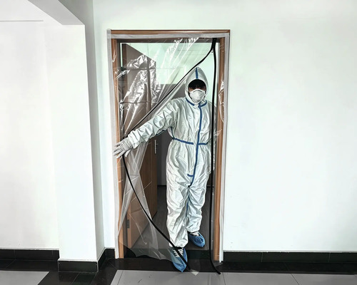 Transparente Staubschutztür mit bogenförmigem Reißverschluss an einem Türrahmen montiert, eine Person im Schutzanzug öffnet die Folie.