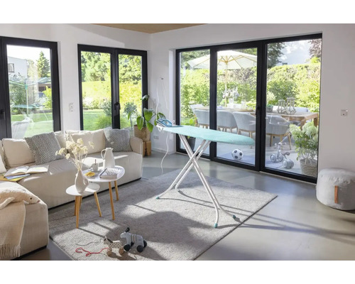 Moderner, lichtdurchfluteter Wohnraum mit aufgestelltem Bügeltisch vor einer großen Fensterfront mit Blick in den Garten.