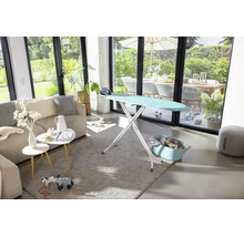 Bügeltisch in einem hellen Wohnzimmer mit Bügeleisen, Wäschekorb, Sofa und Blick auf eine Terrasse.