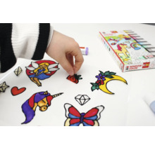 Hand zieht ein selbstgemachtes Window Color Motiv von einer Folie ab, umgeben von bunten Aufklebern wie Einhorn, Fee und Schmetterling.