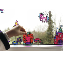 Hand bringt bunte Fensterbilder mit Motiven wie Schloss, Einhorn und Erdbeerhaus auf einer Fensterscheibe an.