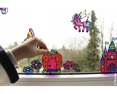 Hand bringt bunte Fensterbilder mit Motiven wie Schloss, Einhorn und Erdbeerhaus auf einer Fensterscheibe an.