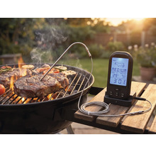 Digitales Grillthermometer mit Fleischsonde misst die Kerntemperatur eines Steaks auf einem Holzkohlegrill in einem hellen Garten.