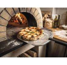 Frisch gebackene Pizza auf einem Pizzaschieber mit Tenneker Logo vor einem brennenden Steinofen in einer rustikalen Küche.