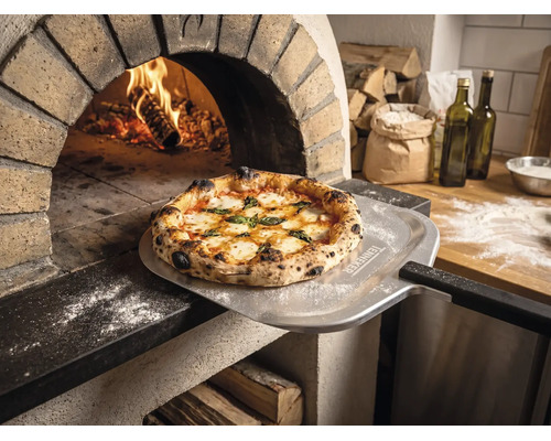 Frisch gebackene Pizza auf einem Pizzaschieber mit Tenneker Logo vor einem brennenden Steinofen in einer rustikalen Küche.