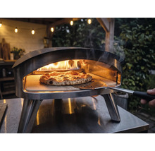 Pizzaofen aus Edelstahl für den Außenbereich mit brennenden Flammen und einer Pizza auf einem Pizzastein, die mit einem Pizzaheber bewegt wird.