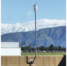 Wetterstation mit weißem Sensor auf einer hohen Metallstange, montiert an einer Mauer vor einer hellen Gebirgskulisse mit schneebedeckten Gipfeln.