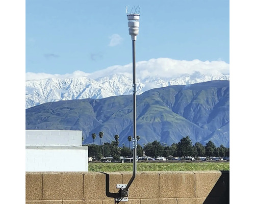 Wetterstation mit weißem Sensor auf einer hohen Metallstange, montiert an einer Mauer vor einer hellen Gebirgskulisse mit schneebedeckten Gipfeln.