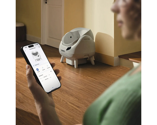 Eine Person steuert eine selbstreinigende Catlink Katzentoilette über eine App auf einem Smartphone in einem hellen Raum.