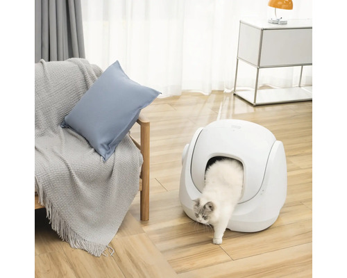 Weiße Katze verlässt eine moderne, selbstreinigende Katzentoilette in einem hellen Wohnraum mit Holzboden und Sessel.