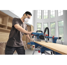 Handwerker mit Schutzausrüstung bedient eine Paneelsäge zum Zuschneiden eines Holzbretts in einer hellen Werkstatt.