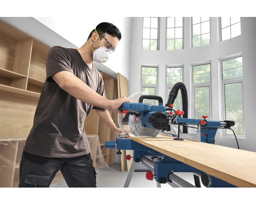 Handwerker mit Schutzausrüstung bedient eine Paneelsäge zum Zuschneiden eines Holzbretts in einer hellen Werkstatt.
