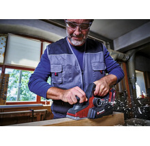 Ein Mann mit Schutzbrille nutzt einen Akkumulator-Hobel mit Einhell Logo zur Bearbeitung eines Holzbalkens in einer hellen Werkstatt.
