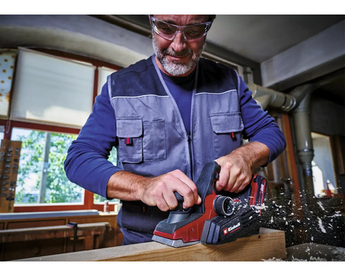 Ein Mann mit Schutzbrille nutzt einen Akkumulator-Hobel mit Einhell Logo zur Bearbeitung eines Holzbalkens in einer hellen Werkstatt.
