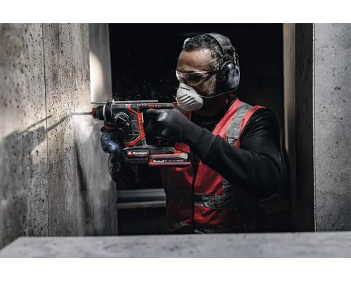 Handwerker bohrt mit Einhell Akku-Bohrhammer in eine Betonwand, trägt Gehörschutz, Schutzbrille und Atemschutzmaske.