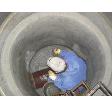 Ein Arbeiter in blauem Schutzanzug dichtet den Boden eines Betonschachts mit Mörtel neben einem Rohr ab.
