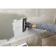 Hand mit Arbeitshandschuh trägt Fliesenkleber mit einer Zahnkelle auf eine verputzte Wand auf.