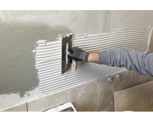 Hand mit Arbeitshandschuh trägt Fliesenkleber mit einer Zahnkelle auf eine verputzte Wand auf.