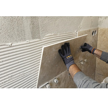 Verlegen einer großformatigen Wandfliese auf Fliesenkleber, Hände in Arbeitshandschuhen mit PCI Logo halten die Fliese an die Wand.