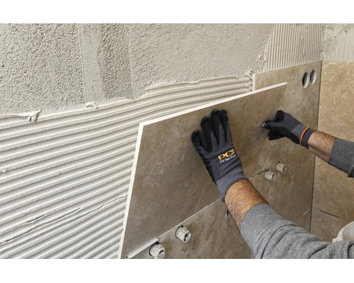 Verlegen einer großformatigen Wandfliese auf Fliesenkleber, Hände in Arbeitshandschuhen mit PCI Logo halten die Fliese an die Wand.