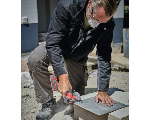 Handwerker mit Schutzbrille und Atemschutzmaske schneidet eine Steinplatte mit einer Akku-Mini-Handkreissäge auf einer Baustelle.