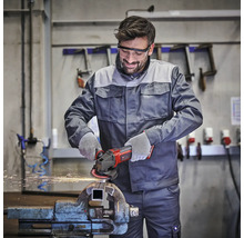 Handwerker nutzt Winkelschleifer an einem Werkstück im Schraubstock, trägt Schutzbrille und Handschuhe in einer hellen, organisierten Werkstatt.