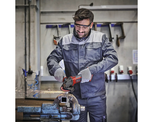 Handwerker nutzt Winkelschleifer an einem Werkstück im Schraubstock, trägt Schutzbrille und Handschuhe in einer hellen, organisierten Werkstatt.
