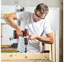 Ein Mann bohrt mit einem Einhell Akku-Bohrschrauber in einen Holzbalken in einer hellen Werkstatt. Er trägt eine Schutzbrille.