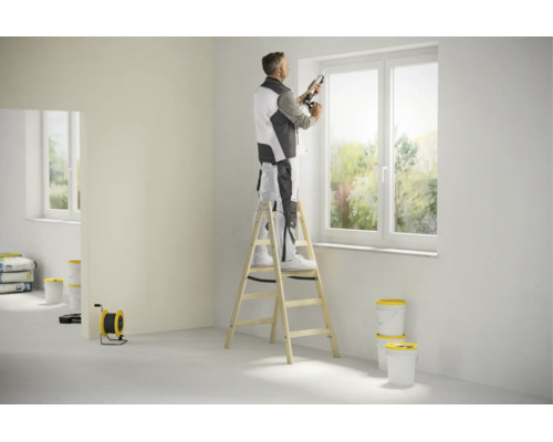 Handwerker steht auf einer Holz-Stufenstehleiter und dichtet ein Fenster mit einer Kartuschenpresse in einem hellen Raum ab.