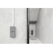 Grauer Zweifach-Wippschalter für die Aufputzmontage an einer grauen Betonwand mit einem weißen Elektroinstallationsrohr.