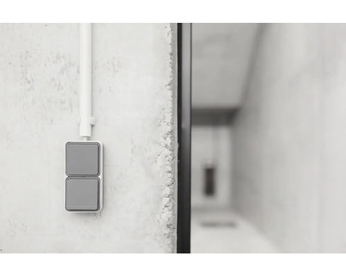 Grauer Zweifach-Wippschalter für die Aufputzmontage an einer grauen Betonwand mit einem weißen Elektroinstallationsrohr.