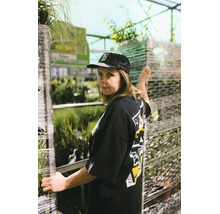 Frau mit schwarzem T-Shirt und Kappe steht in einem Gewächshaus zwischen Pflanzenregalen, die mit Folie umwickelt sind.