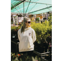 Eine Frau in einem cremefarbenen Hoodie mit Logo steht in einem hellen, organisierten Gartencenter inmitten zahlreicher grüner Pflanzen.