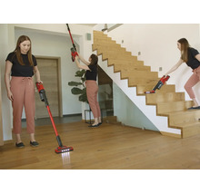 Eine Frau nutzt einen roten Akku-Stielsauger für Holzböden, hohe Wände und Treppen in einem hellen, modernen Wohnraum mit Holztreppe.