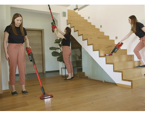 Eine Frau nutzt einen roten Akku-Stielsauger für Holzböden, hohe Wände und Treppen in einem hellen, modernen Wohnraum mit Holztreppe.