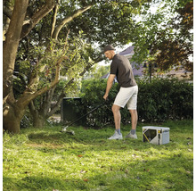 Mann bei der Gartenpflege mit einem elektrischen Rasentrimmer und einer tragbaren Powerstation auf einer Rasenfläche unter Bäumen.